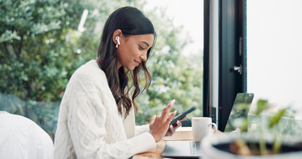 A woman using her phone beside a laptop at a sunlit desk